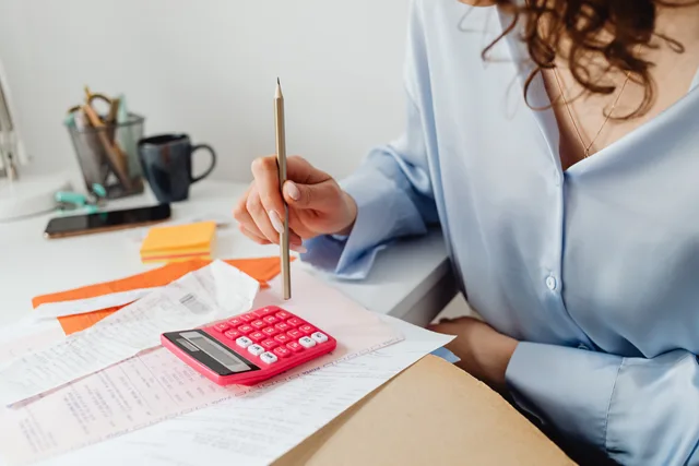 A woman is calculating expenses using a calculator and papers at her desk. Momsredovisning för småföretag från kvitto till deklaration